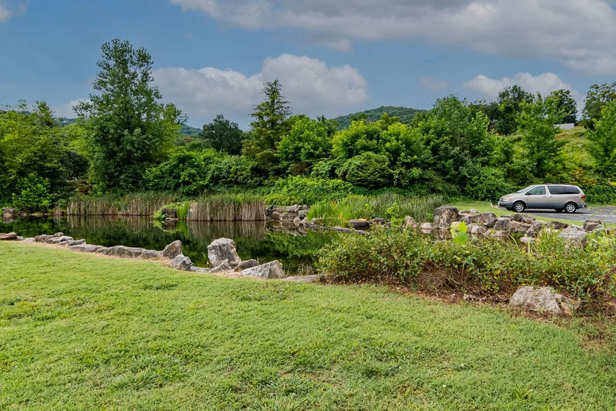 A tranquil pond bordered by rocks and lush landscaping. The scene evokes a peaceful natural setting ideal for picnics, fishing, or a relaxing walk, with a quiet vehicle in the distance suggesting easy access.