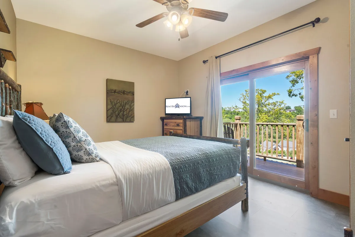 A restful bedroom featuring a cozy bed, a window with a tranquil view, a wall-mounted TV, and tasteful artwork. The décor is peaceful, with soft lighting inviting relaxation.