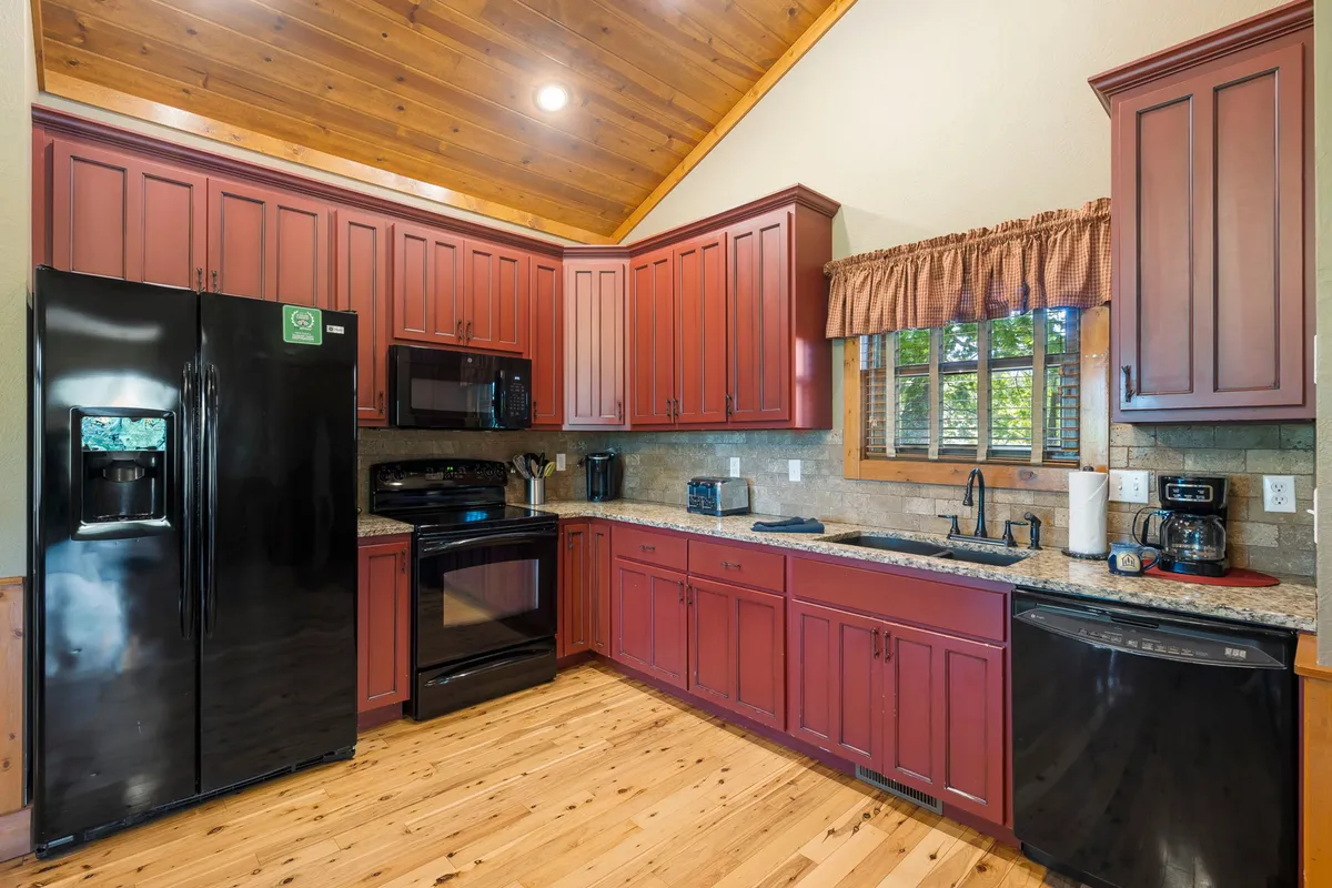 A warm and welcoming kitchen featuring red cabinetry, black appliances, granite countertops, and a wooden floor. Natural light streams in through the window, illuminating the spacious counters and modern appliances, while the layout invites both cooking and socializing.