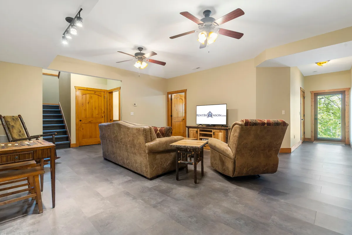 A cozy living room featuring plush sofa seating arranged around a coffee table, a large flat-screen TV mounted above a fireplace, and wooden doors leading to adjoining spaces. The room includes a staircase, suggesting multiple levels and open sightlines, while soft lighting and decor create a welcoming ambiance perfect for relaxing or socializing.
