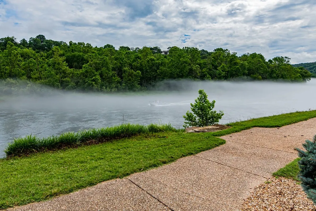 A breathtaking view of the river snaking through lush green trees and shrouded in gentle morning fog. A pathway runs alongside the river, inviting walks, fishing, and peaceful contemplation in nature.