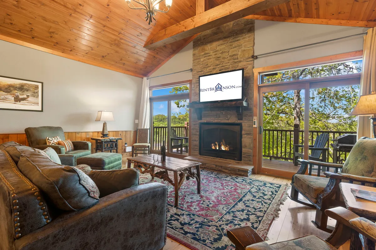 A sunlit living area with a fireplace as the focal point, large windows offering tranquil wooded views, a soft area rug, and a warm wooden ceiling. The space exudes comfort and invites relaxation, with natural light streaming in and furniture arranged for conversation and shared experiences.