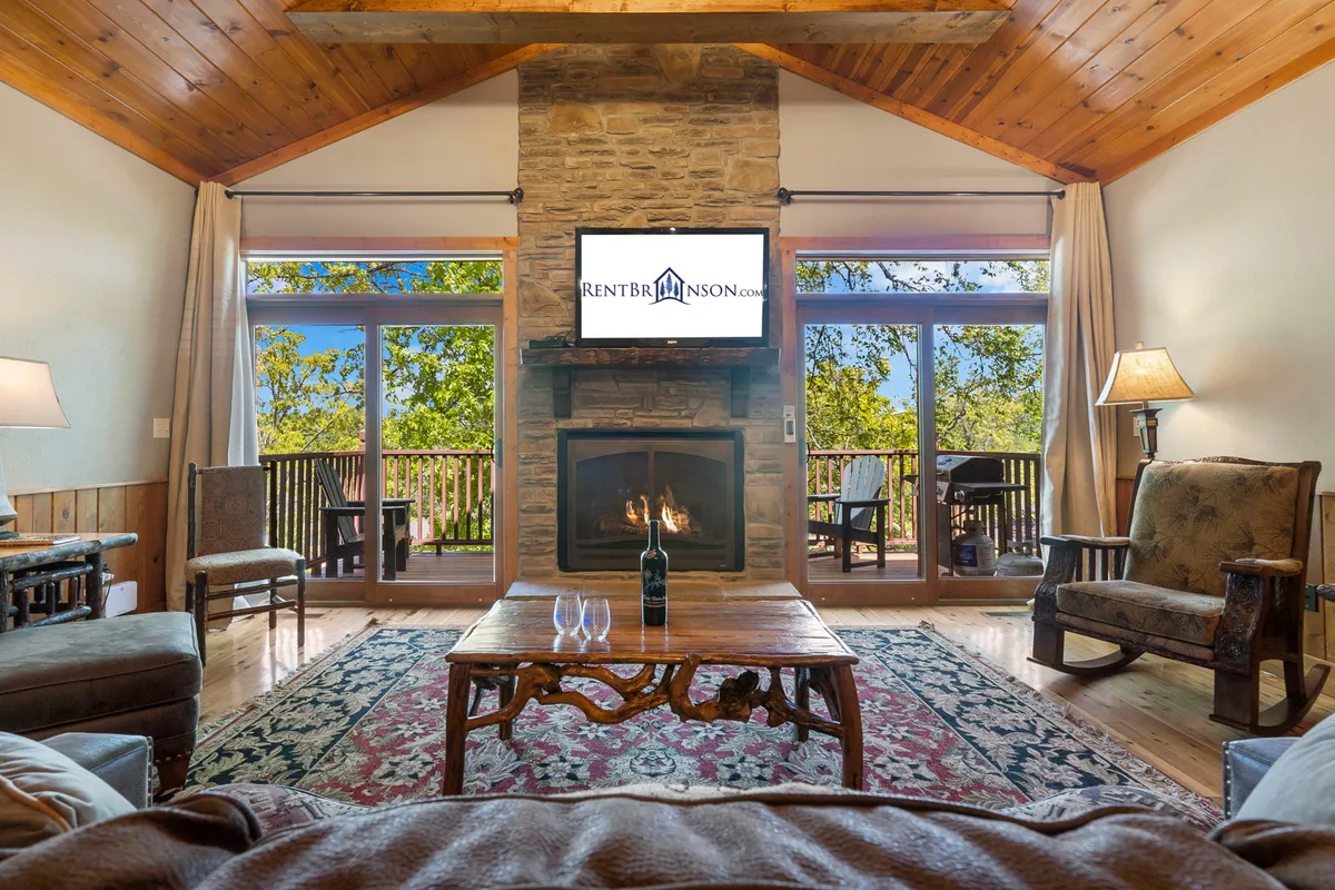 A bright, open living space with a fireplace, coffee table, expansive windows showcasing outdoor greenery, and exposed wooden beams. The seating area encourages conversation and leisure, while the design highlights connectivity with surrounding spaces and the outdoors.
