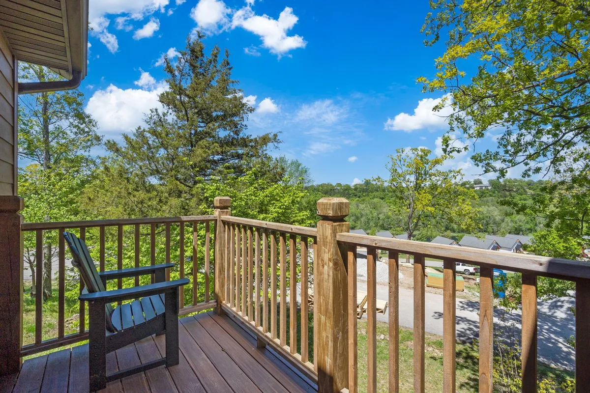A peaceful private patio with a wooden railing, an adirondack chair, and lush greenery, framed by a blue sky with clouds. The setting invites guests to unwind, enjoy nature, or relax with a book after a day of activities.