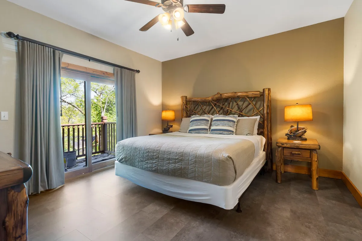 A serene bedroom featuring a wooden bed frame with plush, decorative pillows and nightstands with lamps. The large window offers a beautiful view and fills the space with natural light, creating a cozy and restful atmosphere.